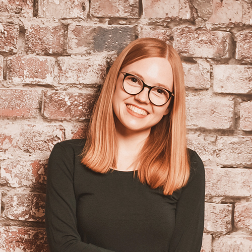 eine Frau mit roten Haaren und Brollen sitzt lächelnd vor einer Backsteinwand.