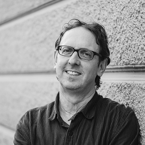 Schwarz-Weiß-Portraits eines Mannes mit Brille, der lächelnd vor einer Wand steht.