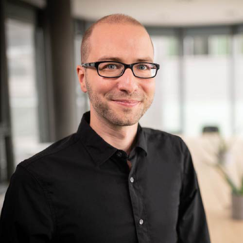 Ein Dozent mit schwarzem Hemd und Brille lächelt in die Kamera