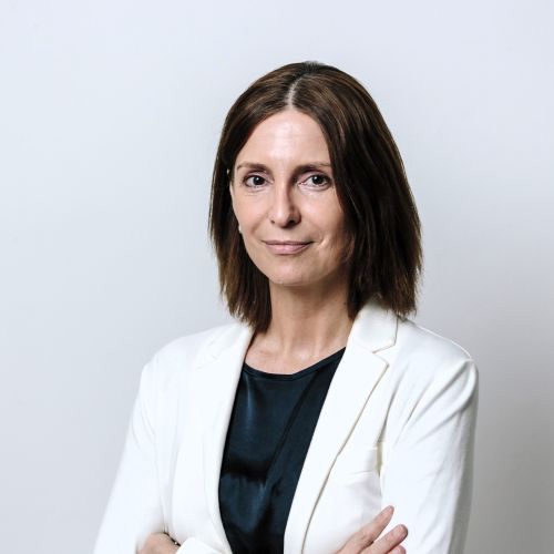 Dozentin mit mittellangen braunen Haaren bekleidet mit einem schwarzen T-Shirt und einem weißen Blazer